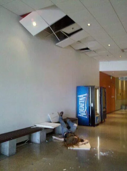 A person is lying on the floor next to a bench with a laptop, surrounded by scattered papers, while the ceiling tiles above…