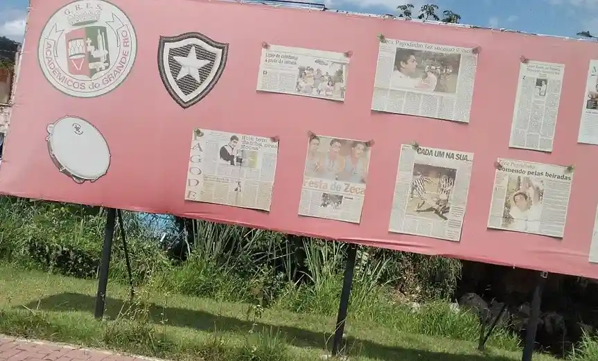 A pink outdoor display board features the logo of 'Acadêmicos do Grande Rio' and a black-and-white star shield. Several…