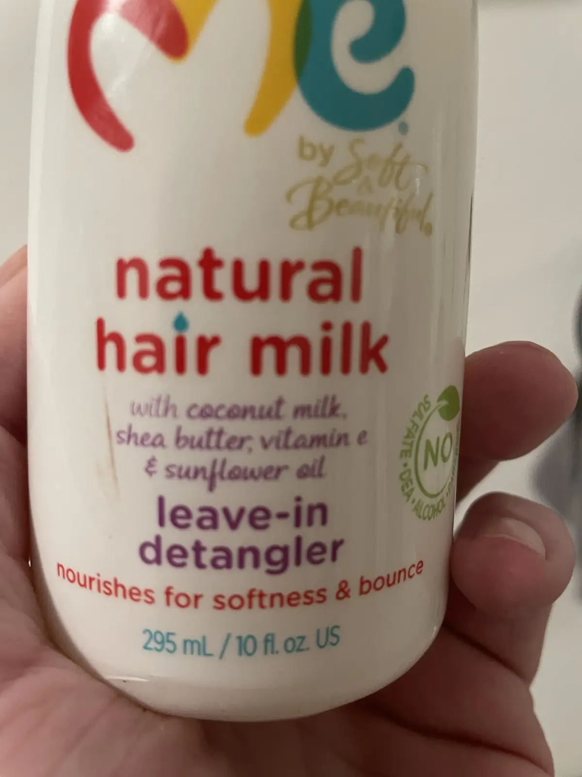 A hand holds a white bottle of 'Me by Soft Beautiful' natural hair milk, which is labeled as a leave-in detangler with…