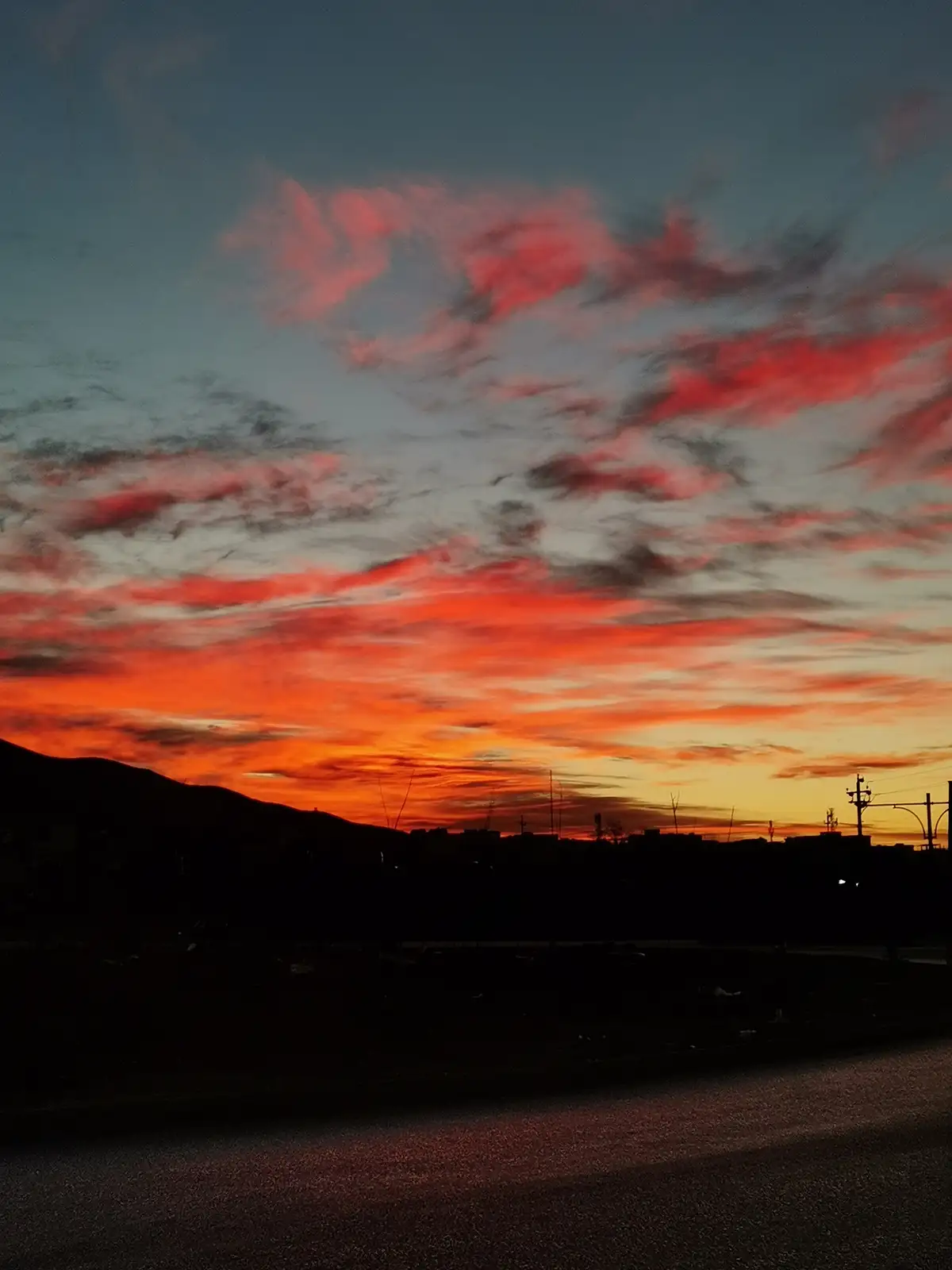 A dramatic sunset with fiery orange and red clouds against a dark blue sky, silhouetting a distant mountain range and urban…