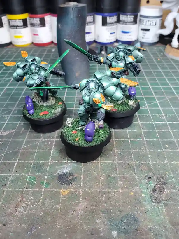 Three painted miniature sci-fi warriors in green armor stand on grassy bases with purple mushrooms, surrounded by paint…