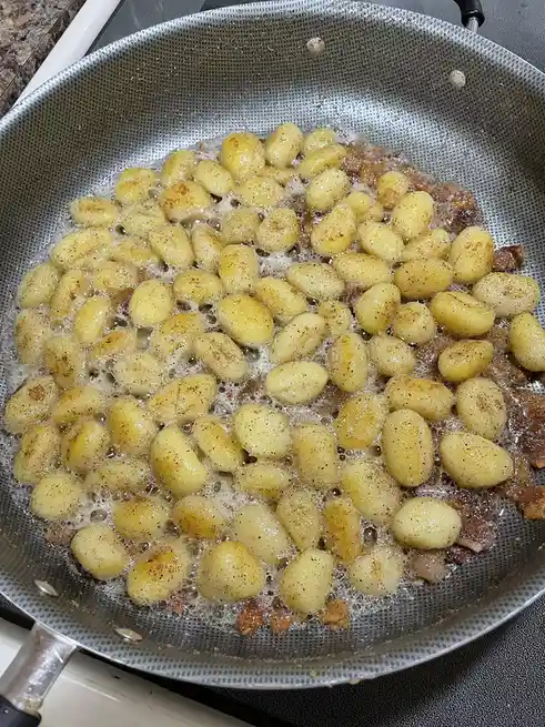 A close-up photo shows small, round, pale-yellow gnocchi sizzling in a silver perforated frying pan with bubbling oil and…