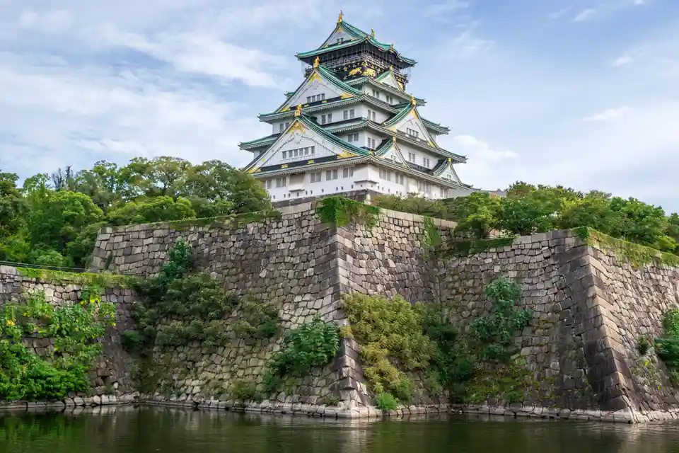 A photograph of Osaka Castle, a multi-tiered Japanese castle with white walls and green roofs, sits atop a massive stone…