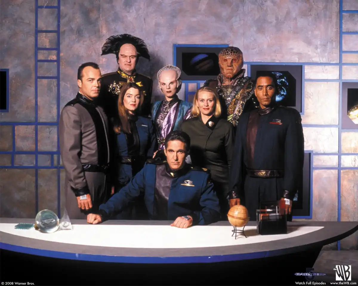 A promotional photo of the cast of 'Babylon 5' posed in their futuristic uniforms around a curved command desk, with a…