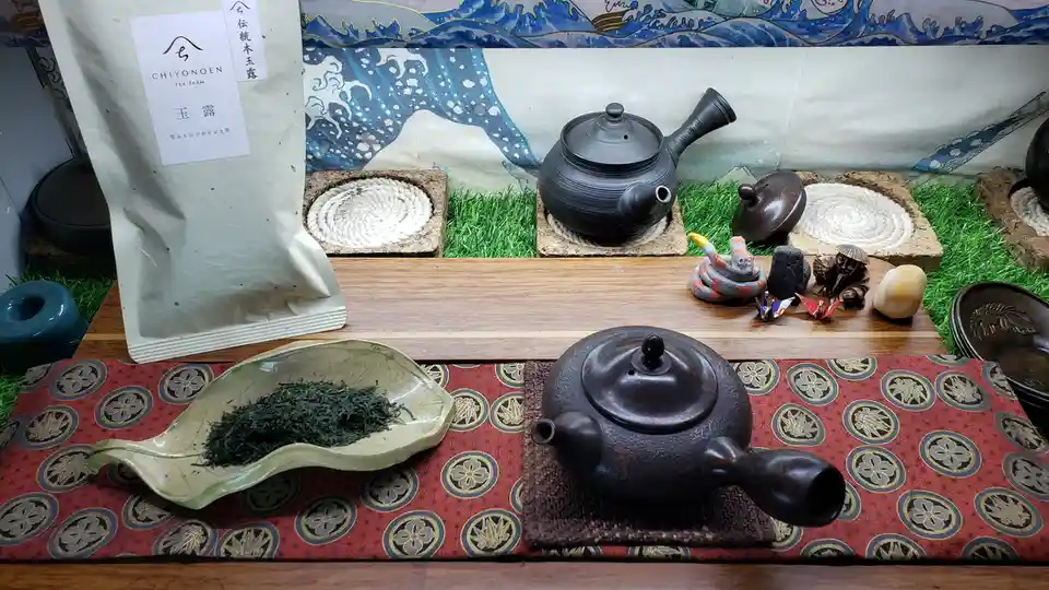 a staged tea setup featuring a large bag of 'Chiyonoen Tea Farm' green tea, a leaf-shaped ceramic dish filled with loose tea…