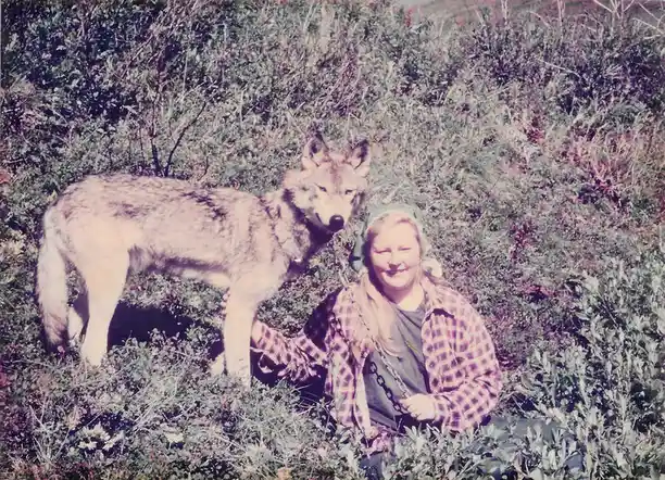 A vintage photo shows a woman in a plaid shirt and headscarf kneeling in a field of low bushes, smiling while holding a…