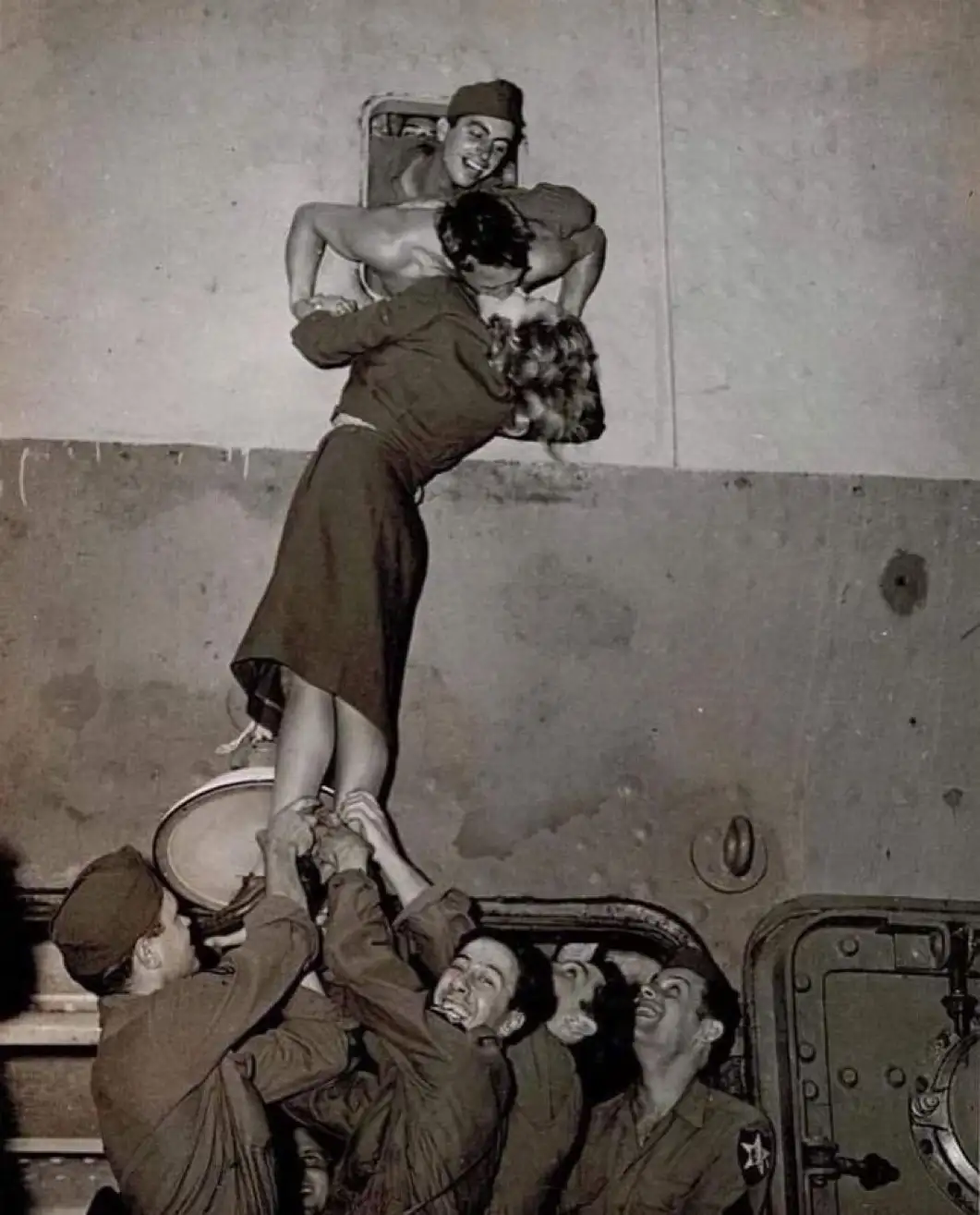 A black-and-white historical photo shows a shirtless man being lifted by several smiling soldiers inside a military vehicle…