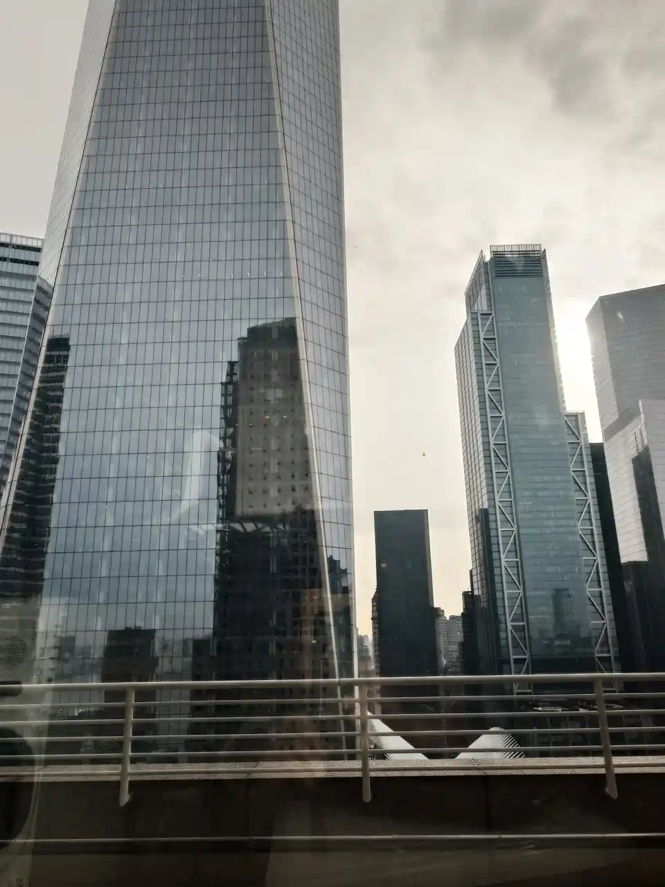 A photo of a modern city skyline with tall glass skyscrapers under an overcast sky, viewed from a balcony or window with a…