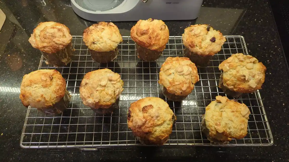 A photo shows nine golden-brown muffins cooling on a wire rack on a dark countertop, some topped with almonds and dried…