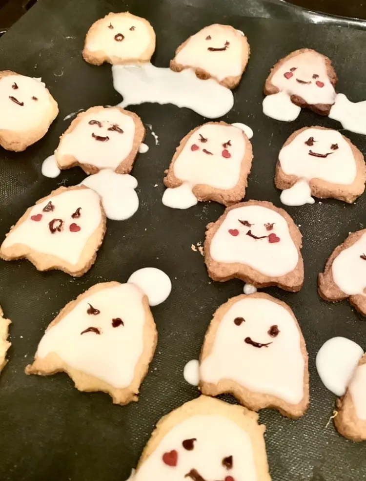 A tray of ghost-shaped sugar cookies decorated with white icing and chocolate facial expressions, some with hearts for…