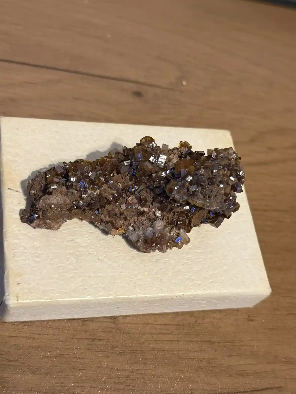 A close-up photo shows a rough, brownish mineral specimen with many small, shiny, reflective crystals embedded in it…