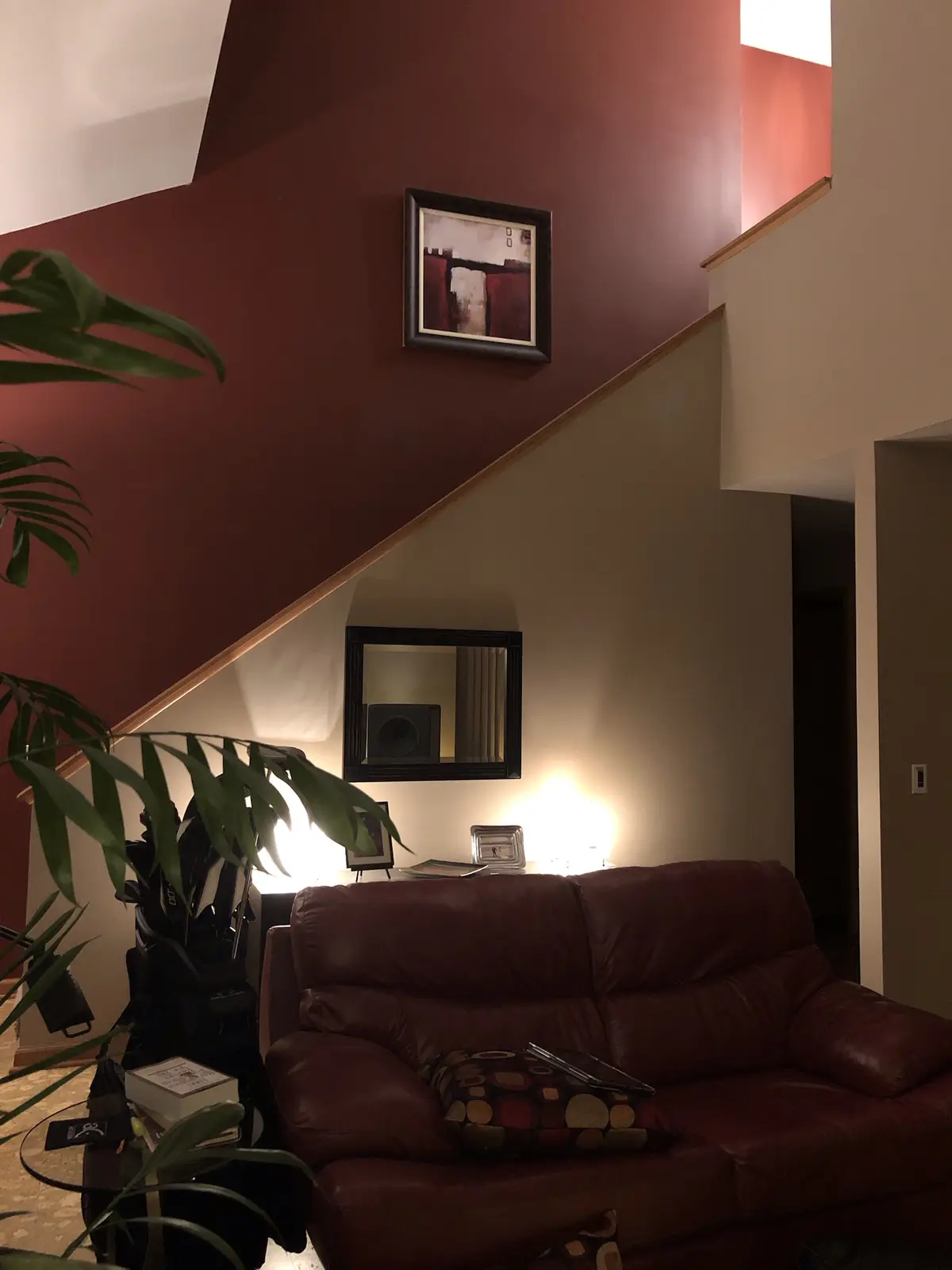 A cozy living room with a red leather couch, a large green plant, and a staircase with a wooden railing. The walls are…