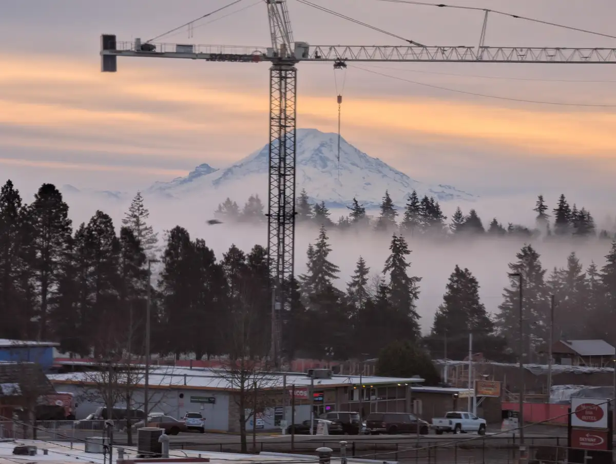 A photo of a construction crane towering over a foggy urban landscape with a snow-capped mountain in the background at…
