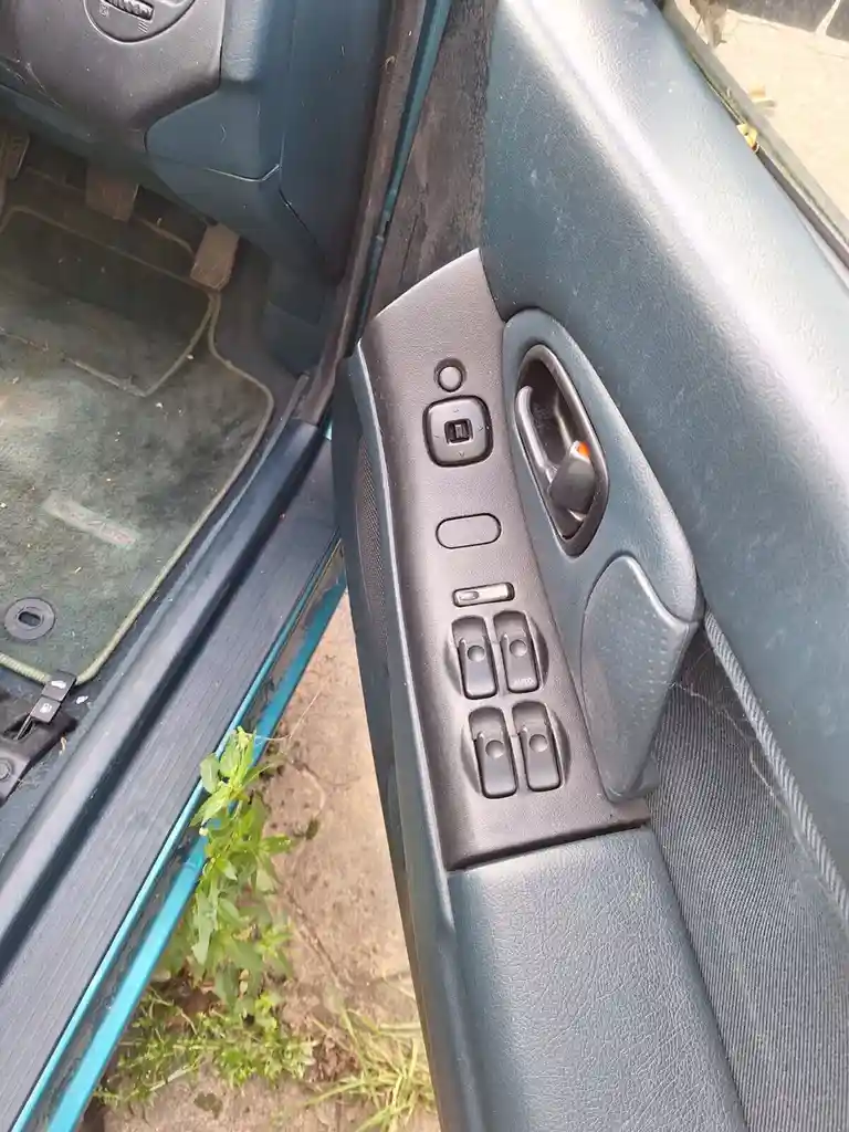 A close-up photo of a car's interior door panel, showing worn black plastic controls for windows and locks, with a green…