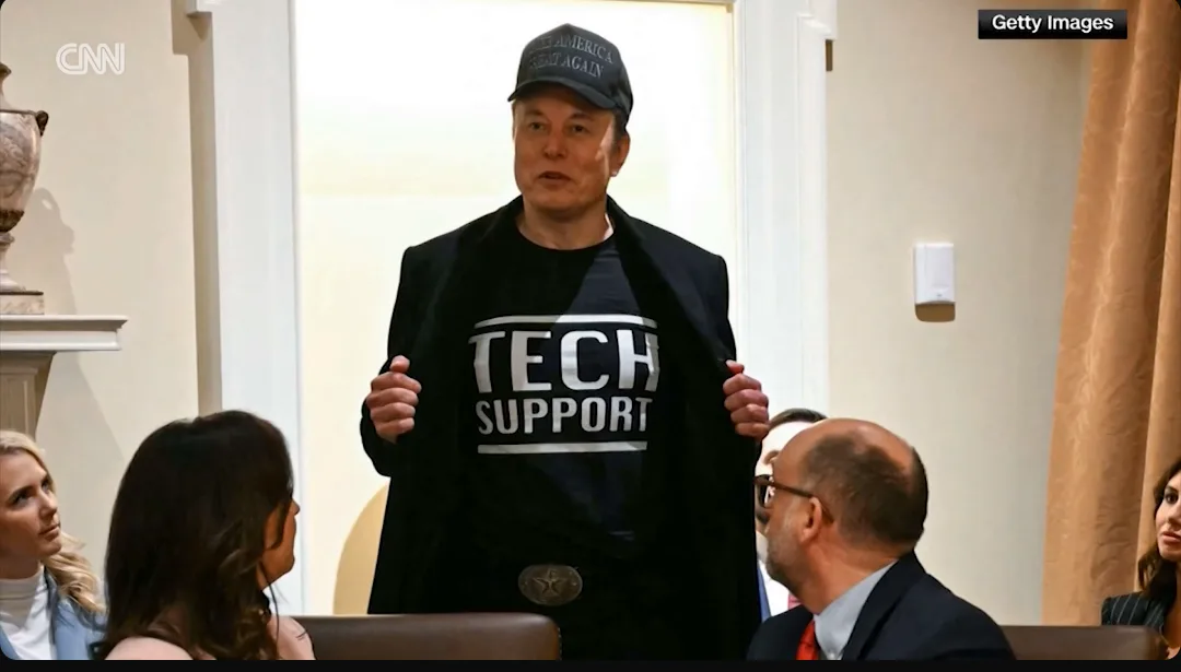 elon musk stands in a room wearing a black jacket over a t-shirt that reads 'TECH SUPPORT' in bold white letters, while…