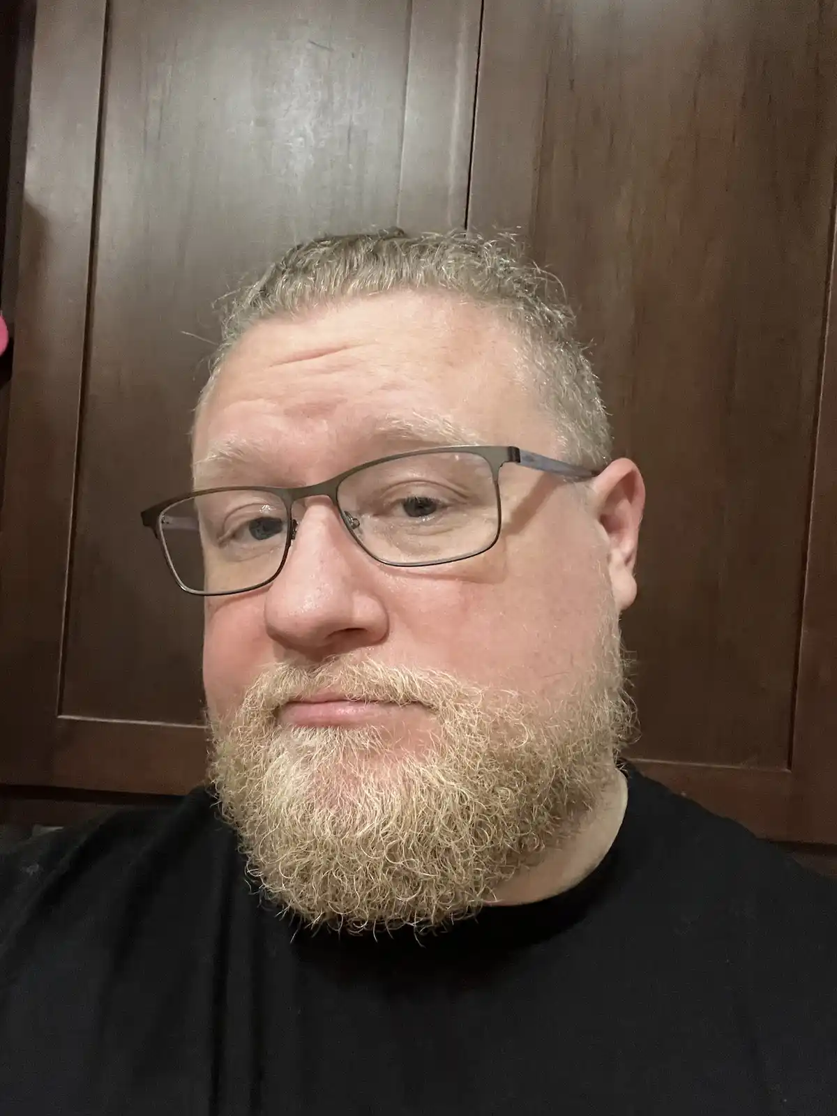 A close-up selfie of a middle-aged man with glasses and a full, curly, light-colored beard, wearing a black shirt against a…