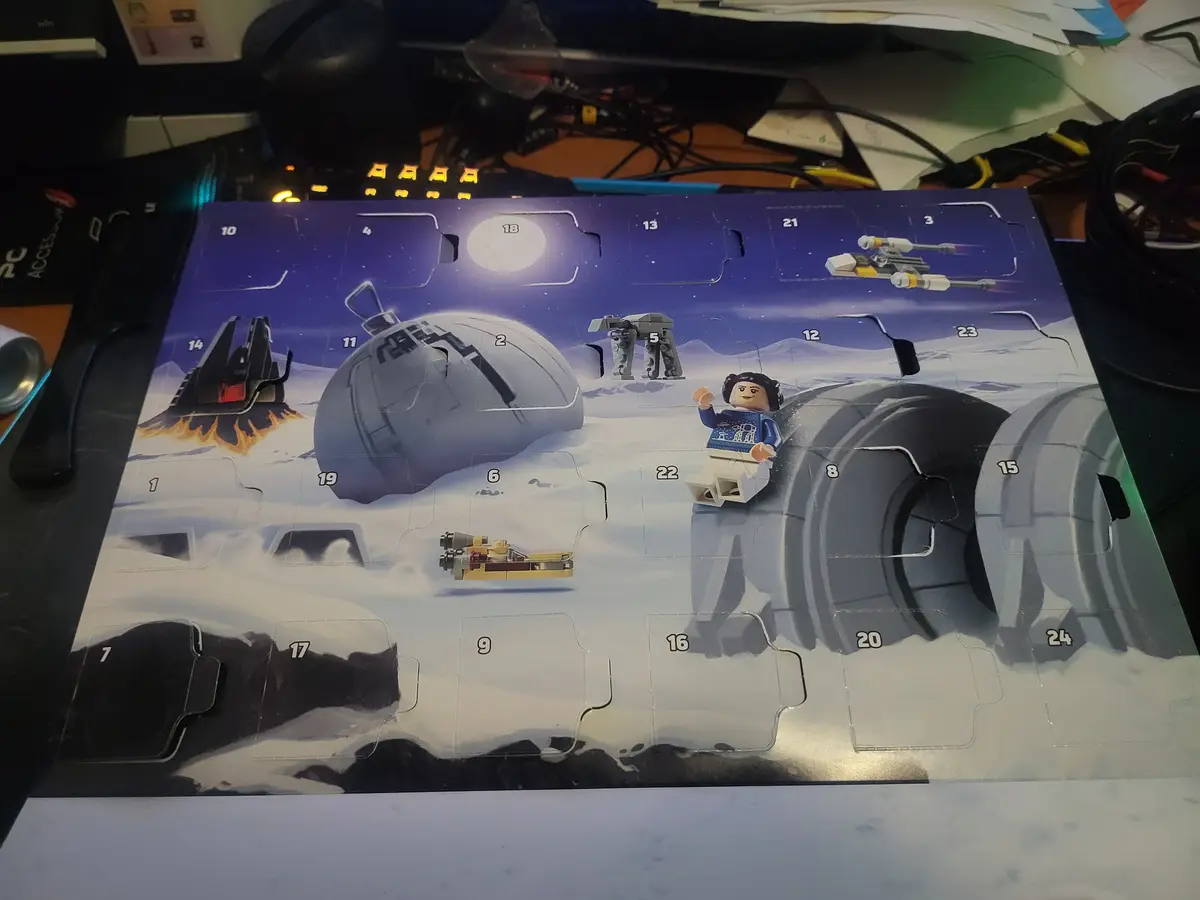 A Lego Star Wars Advent Calendar laid out on a cluttered desk, featuring a snowy Hoth scene with numbered flaps. Several…