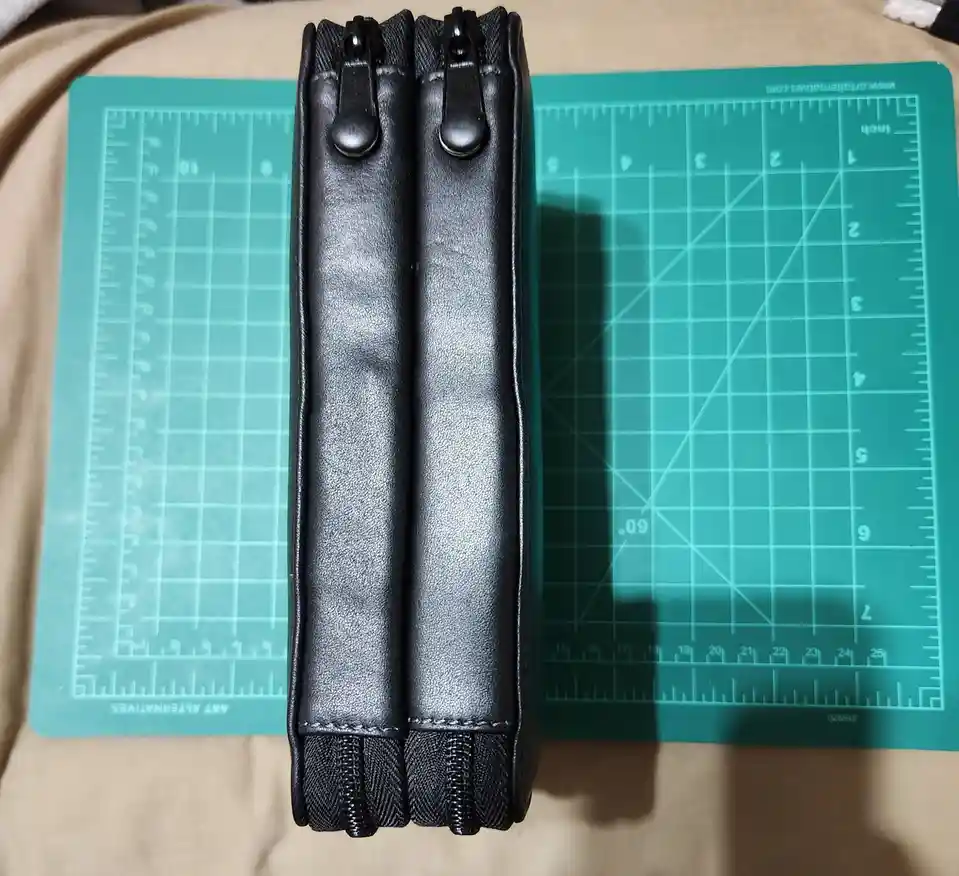 A black leather-like pen case with two parallel zippers sits upright on a green self-healing cutting mat with grid lines and…