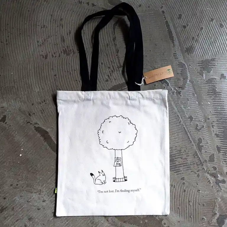 A white tote bag with black straps lies on a textured gray surface. It features a black line drawing of a cat sitting near a…