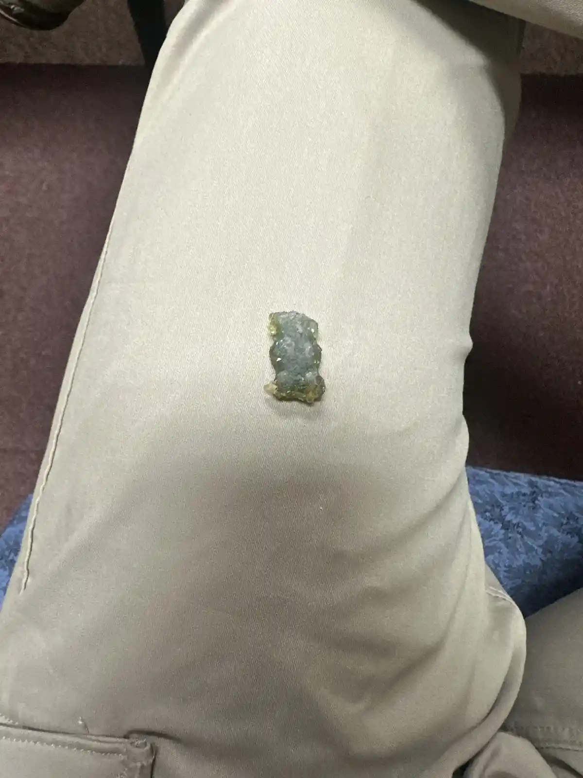A small, translucent, greenish-yellow gummy bear-shaped candy rests on the thigh of someone wearing light beige pants. The…