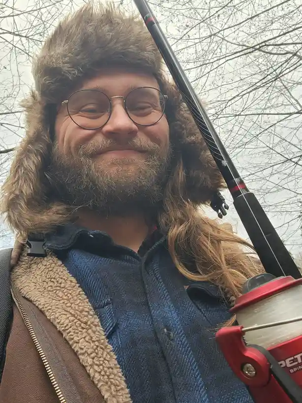 A man with a beard, round glasses, and a furry winter hat smiles at the camera while holding a fishing rod with a red reel…