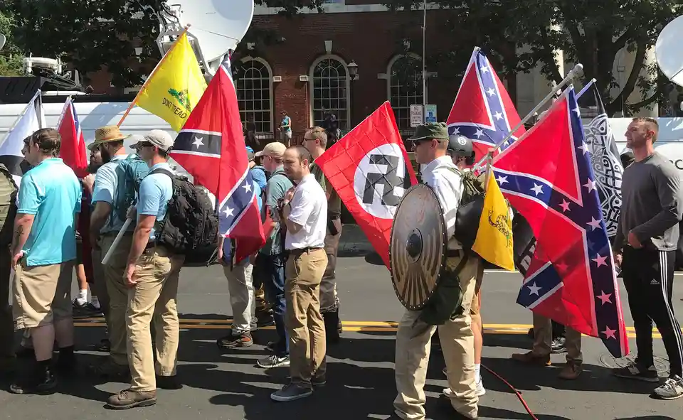 A group of men stand in a street, holding various flags including the Confederate flag, a yellow 'Don't Tread On Me' flag…
