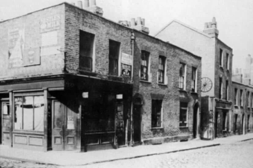 A black-and-white historical photo shows a row of brick buildings on a street corner, with faded signs and a cobblestone…