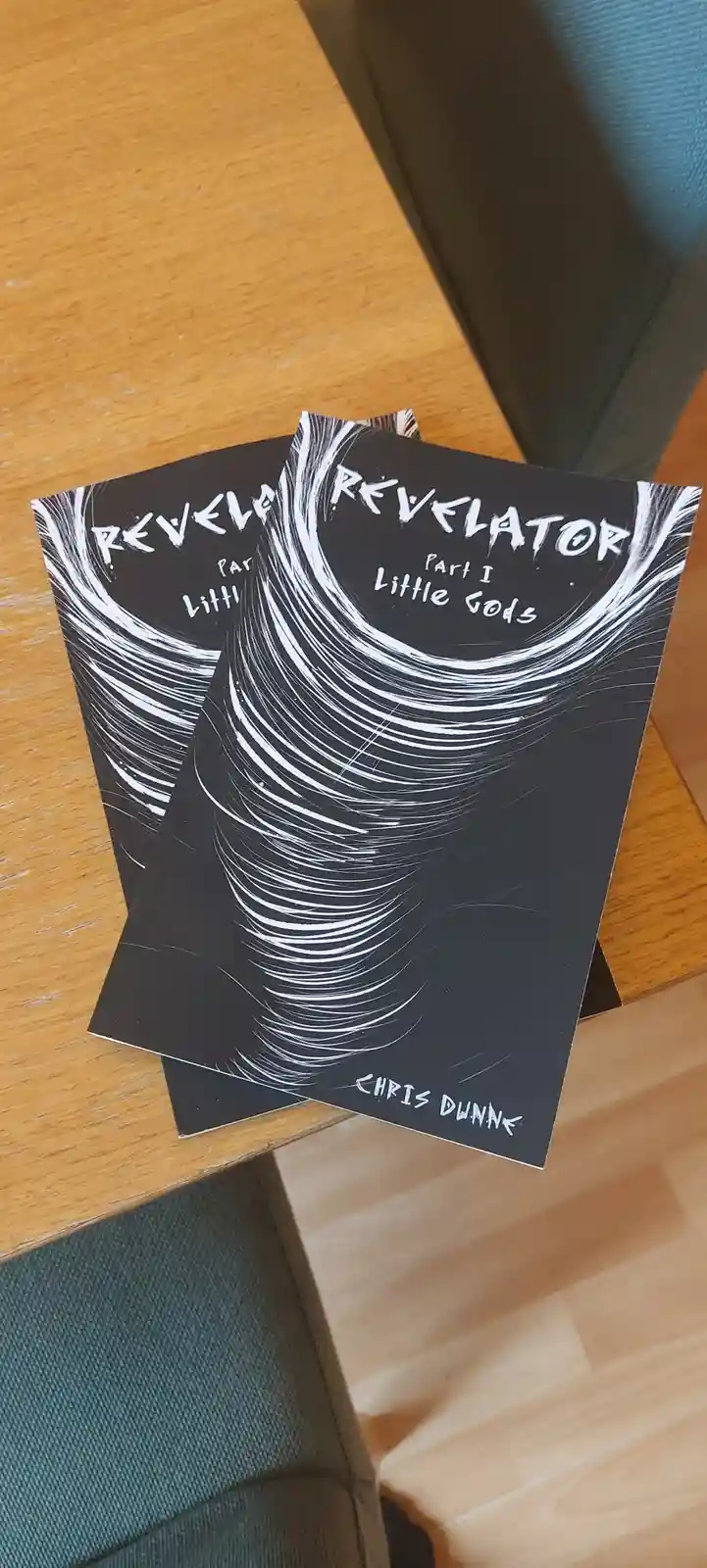 Two identical black-and-white booklets titled 'REVELATOR Part I Little Gods' by Chris Dunne are stacked on a wooden table…