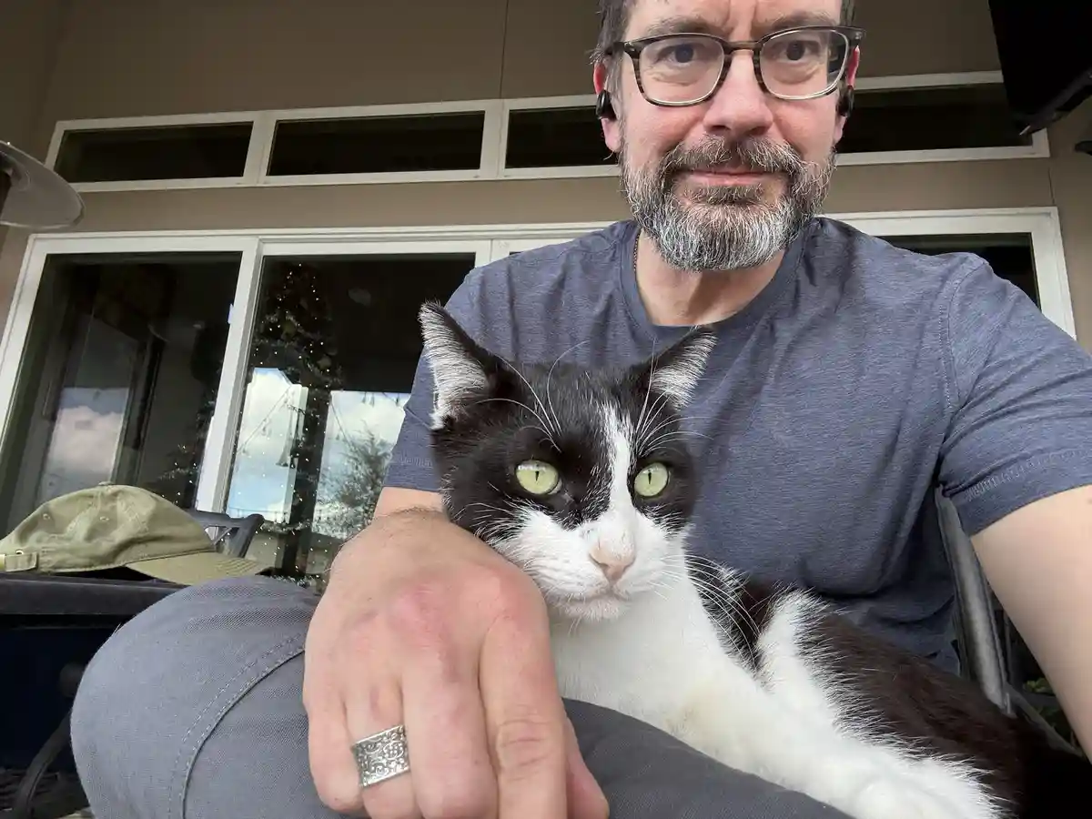 A man with glasses and a beard, wearing a gray t-shirt and earbuds, sits holding a black-and-white cat with striking green…