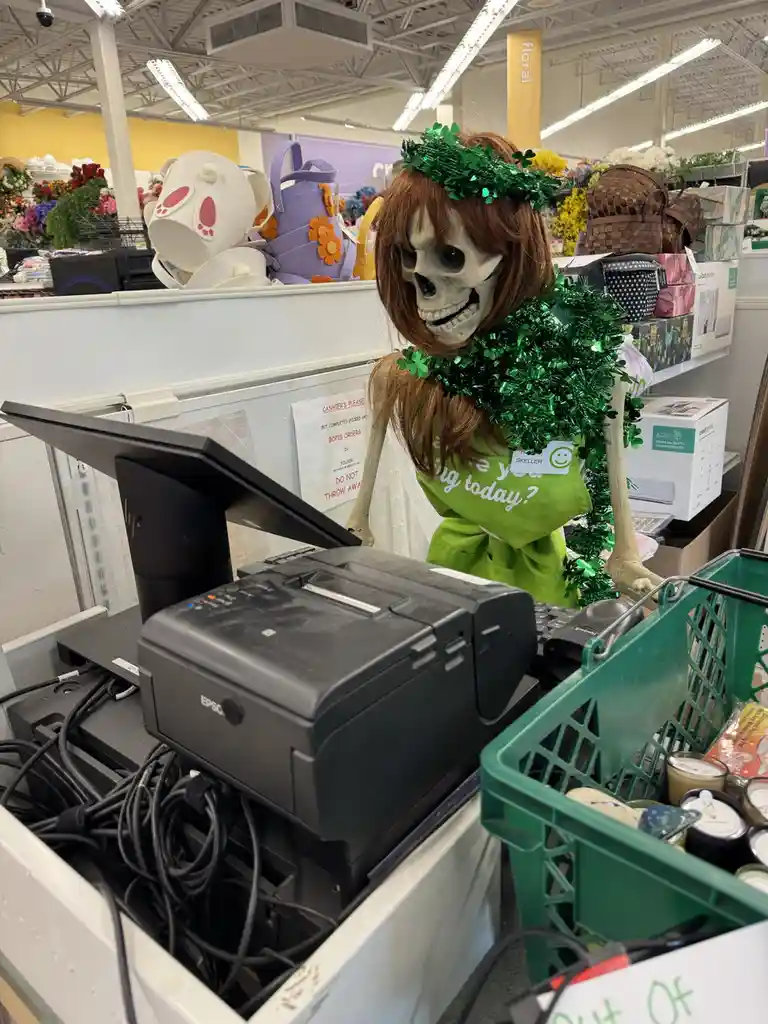 A skeleton dressed as a cashier in a store, wearing a green wig, green tinsel garland, and a green apron with the text 'are…