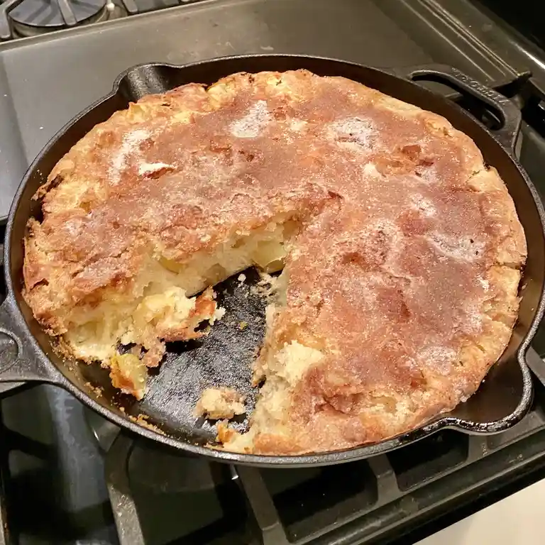 A golden-brown baked dish, resembling a cake or casserole, sits in a black cast-iron skillet on a stovetop. A large, jagged…