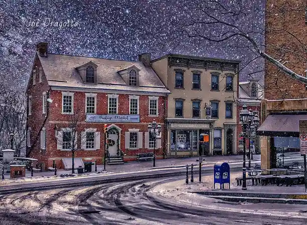 A snowy street scene with brick buildings labeled 'Vintage Market' and 'Visitor Center', a blue mailbox, and tire tracks in…