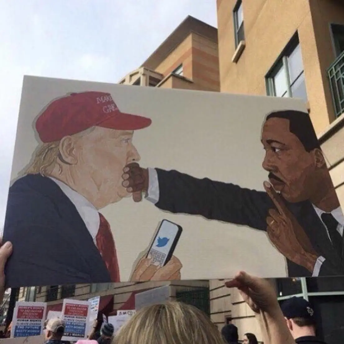 A protest sign depicts a painted scene where Martin Luther King Jr. is shown silencing Donald Trump by placing his finger…
