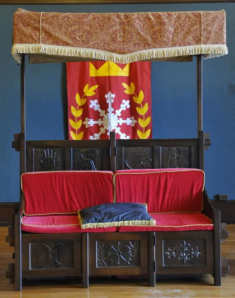 a wooden throne with red cushions and a canopy, positioned in front of a red banner with a white cross and yellow laurel…