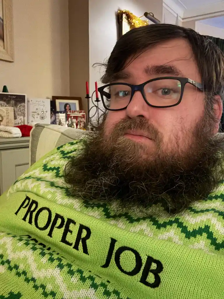 A man with a full beard and glasses wears a bright green, patterned sweater with the words 'PROPER JOB' knitted across the…