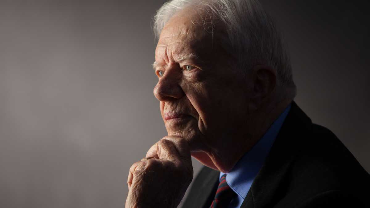 a close-up portrait of an elderly white man with white hair, wearing a dark suit, light blue shirt, and patterned tie…