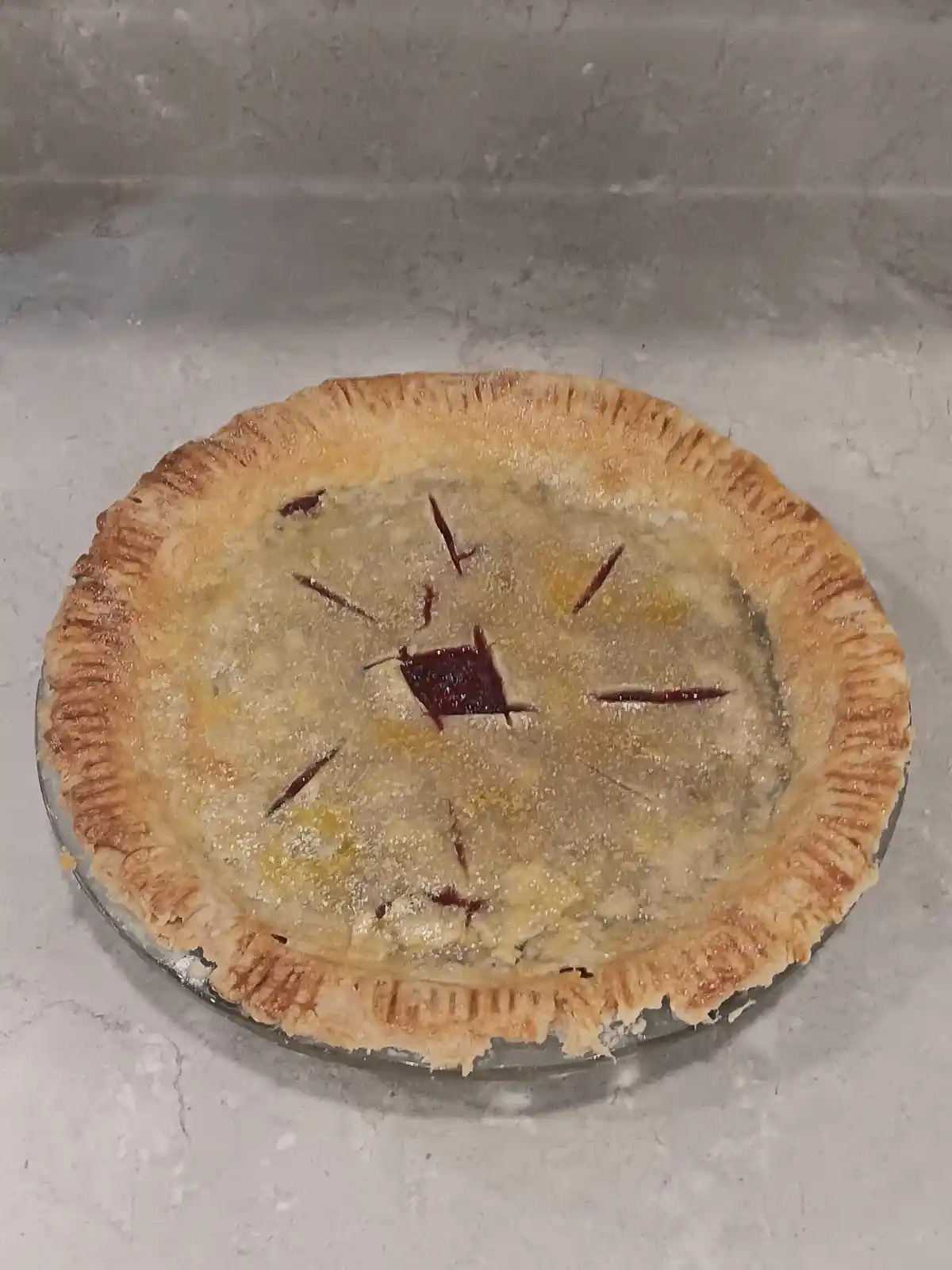 A baked pie with a golden-brown crust sits on a glass dish atop a light gray countertop. The pie’s top has several slits cut…