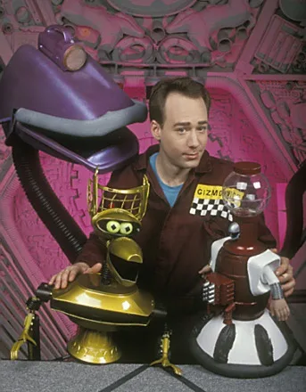 A man in a brown jumpsuit with a 'GIZMOS' patch stands between two robots, one gold with a large purple lamp head and the…