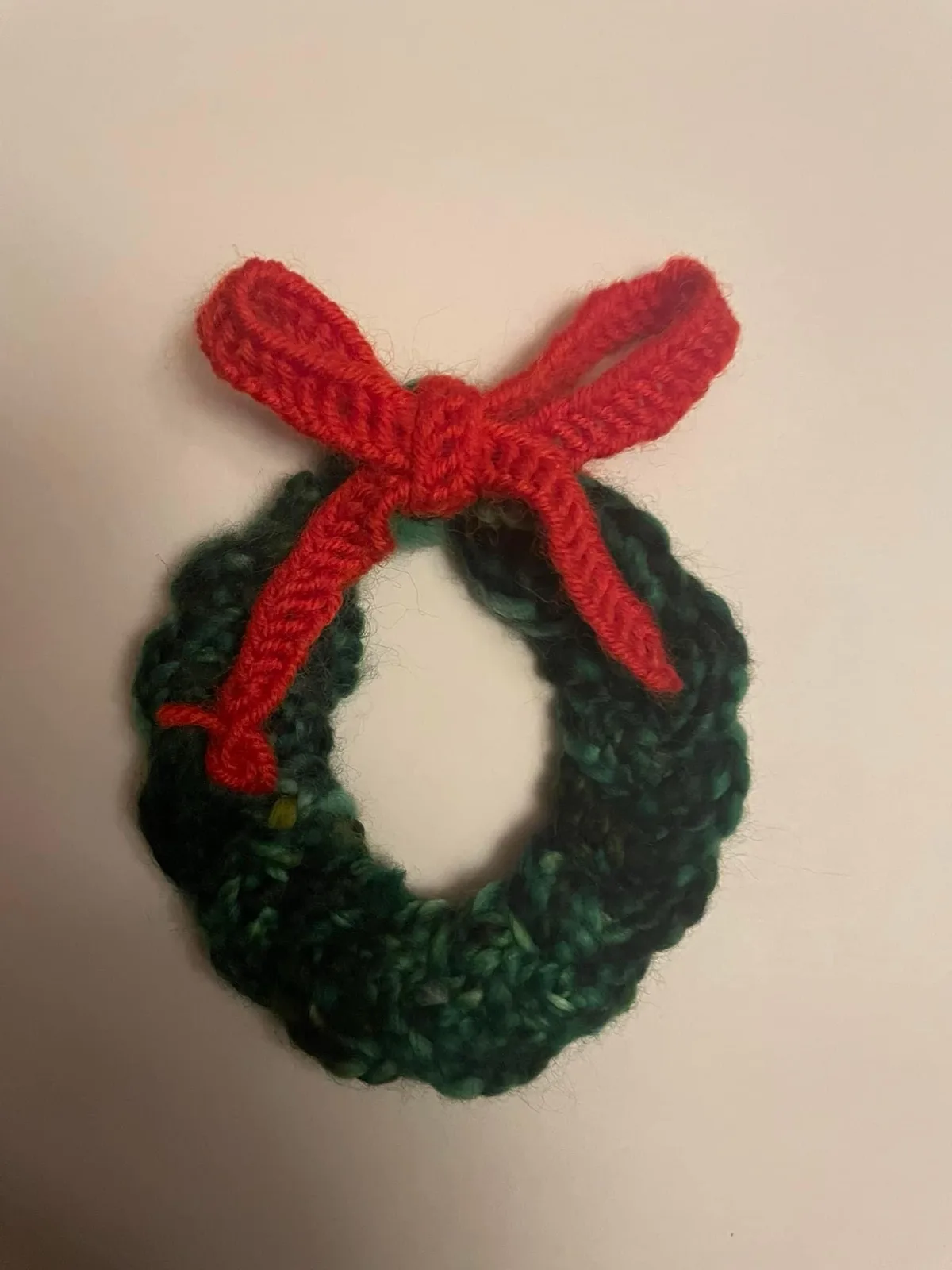 A small, hand-crocheted green Christmas wreath with a red bow sits on a plain white surface. The text overlaid on the image…