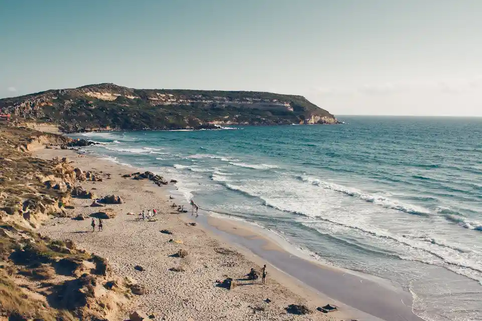 A wide-angle photo of a sunny beach with gentle waves, scattered people enjoying the shore, and a rocky headland in the…