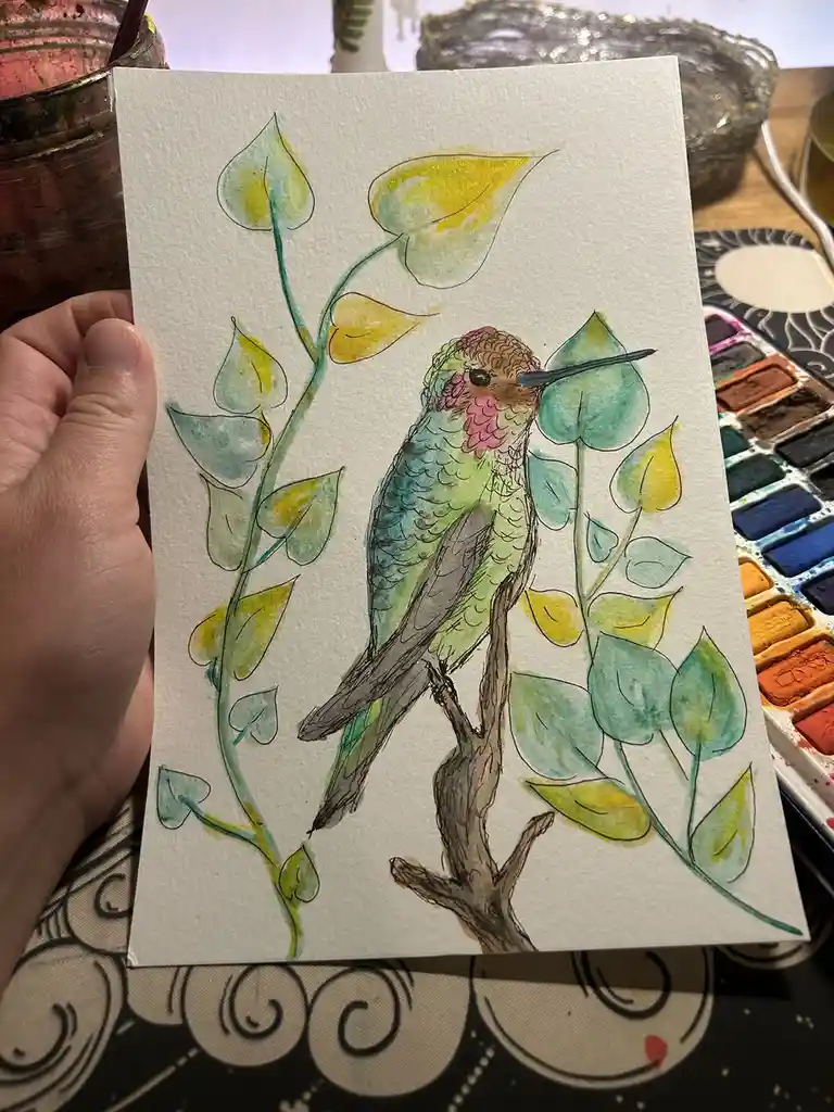 A hand holds up a watercolor painting of a hummingbird perched on a branch surrounded by green and yellow leaves. The bird's…