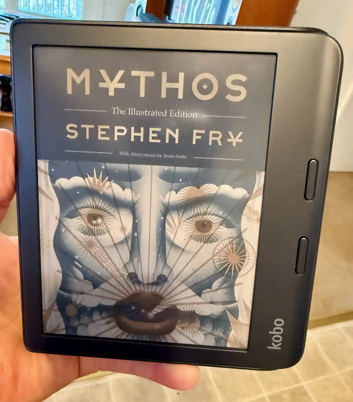 A hand holds a black Kobo e-reader displaying the cover of Stephen Fry's 'Mythos: The Illustrated Edition,' which features a…