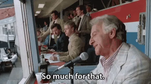 A man in a light-colored suit sits at a desk with a microphone in a broadcast booth, looking toward an audience. The scene…