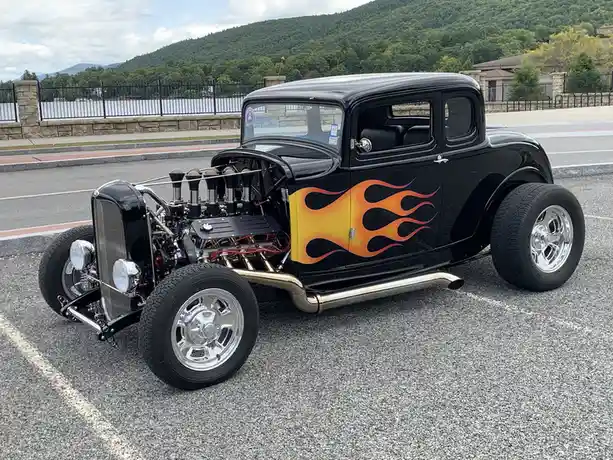 A black classic hot rod with a large exposed engine, chrome wheels, and orange flame decals on its side is parked in a…
