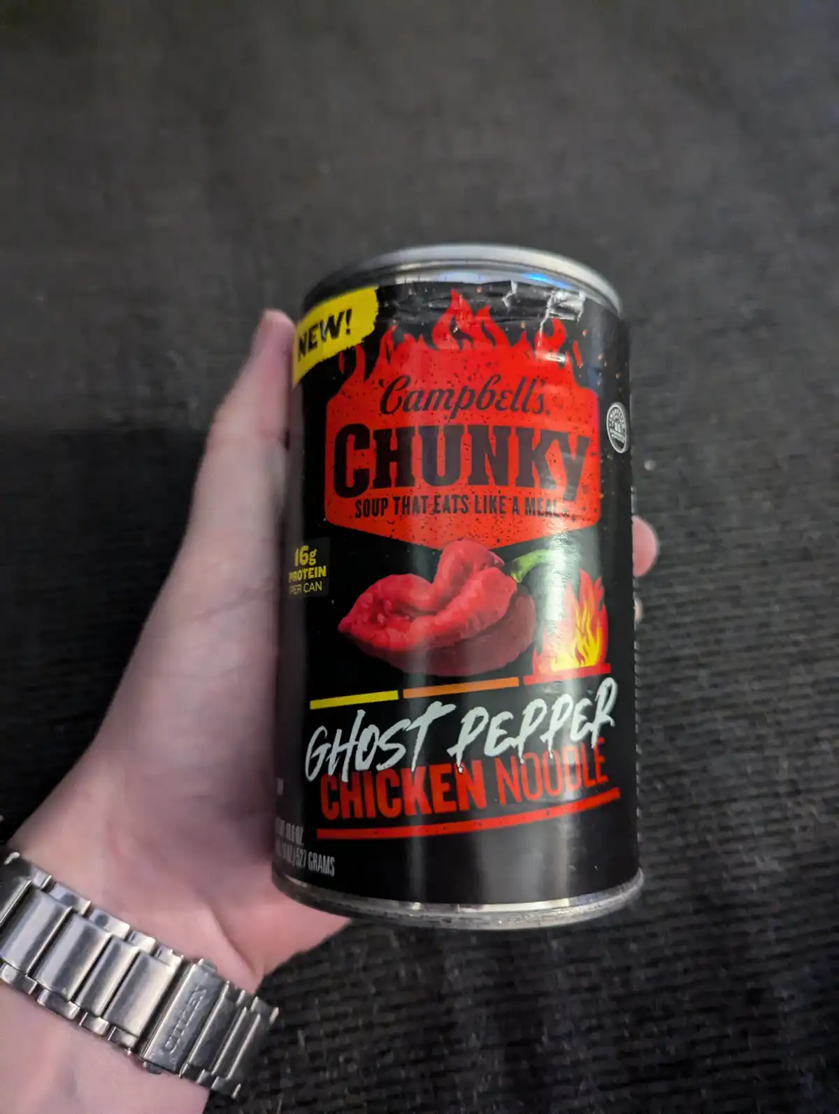 A hand holds a can of Campbell's Chunky soup labeled 'Ghost Pepper Chicken Noodle' against a dark textured background. The…