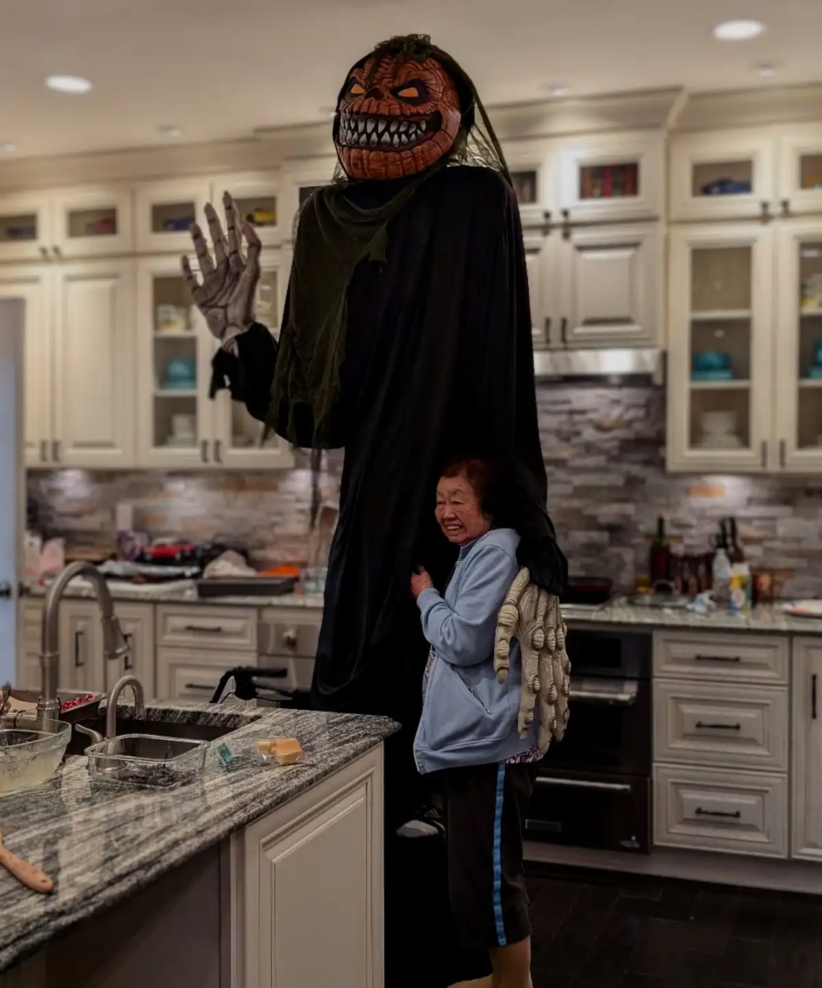 A person in a tall, scary pumpkin-headed costume stands in a modern kitchen, waving one hand while a small elderly woman…
