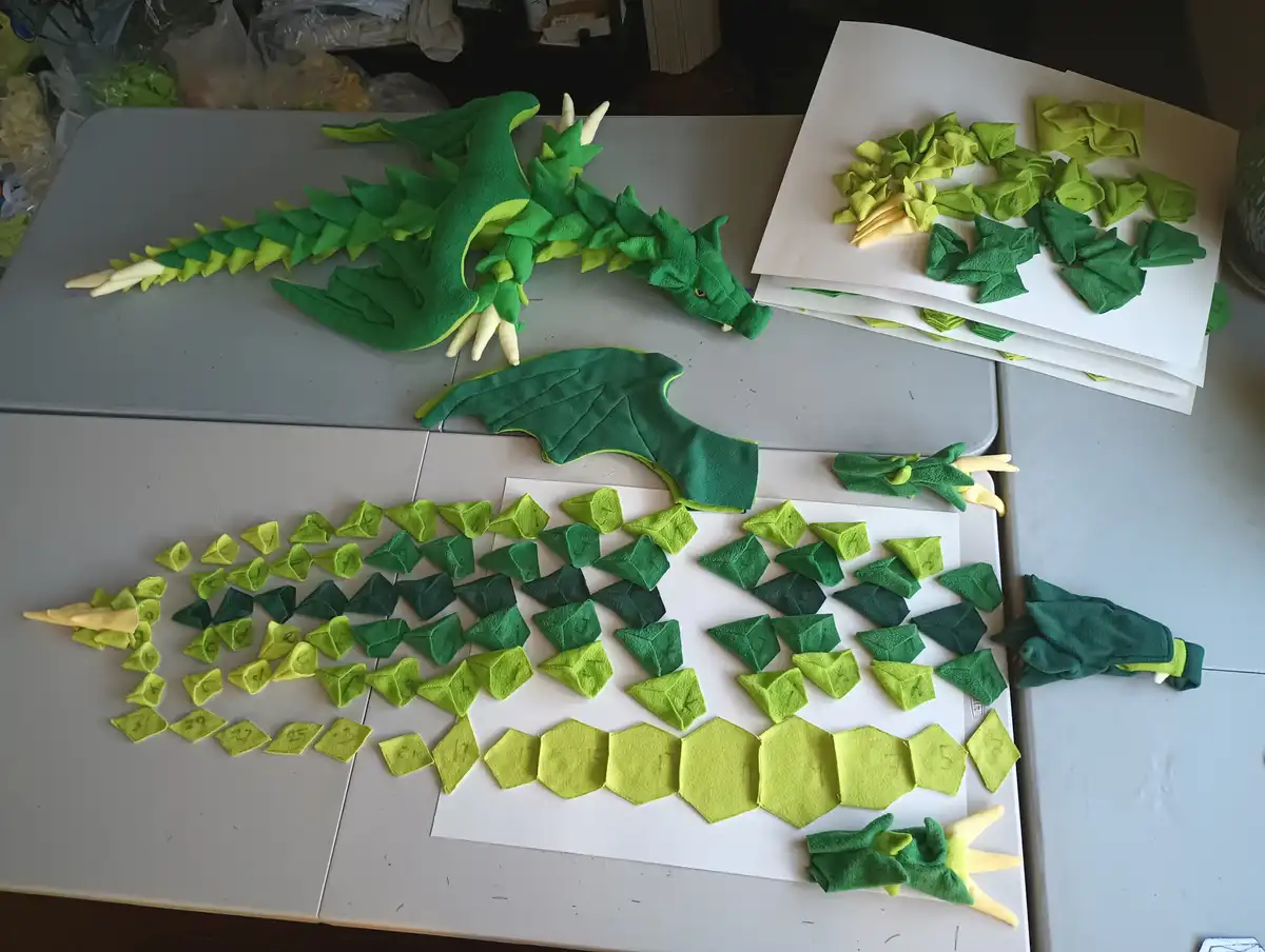 A green felt dragon plush is shown assembled on a table next to piles of its disassembled felt pieces, which are shaped like…