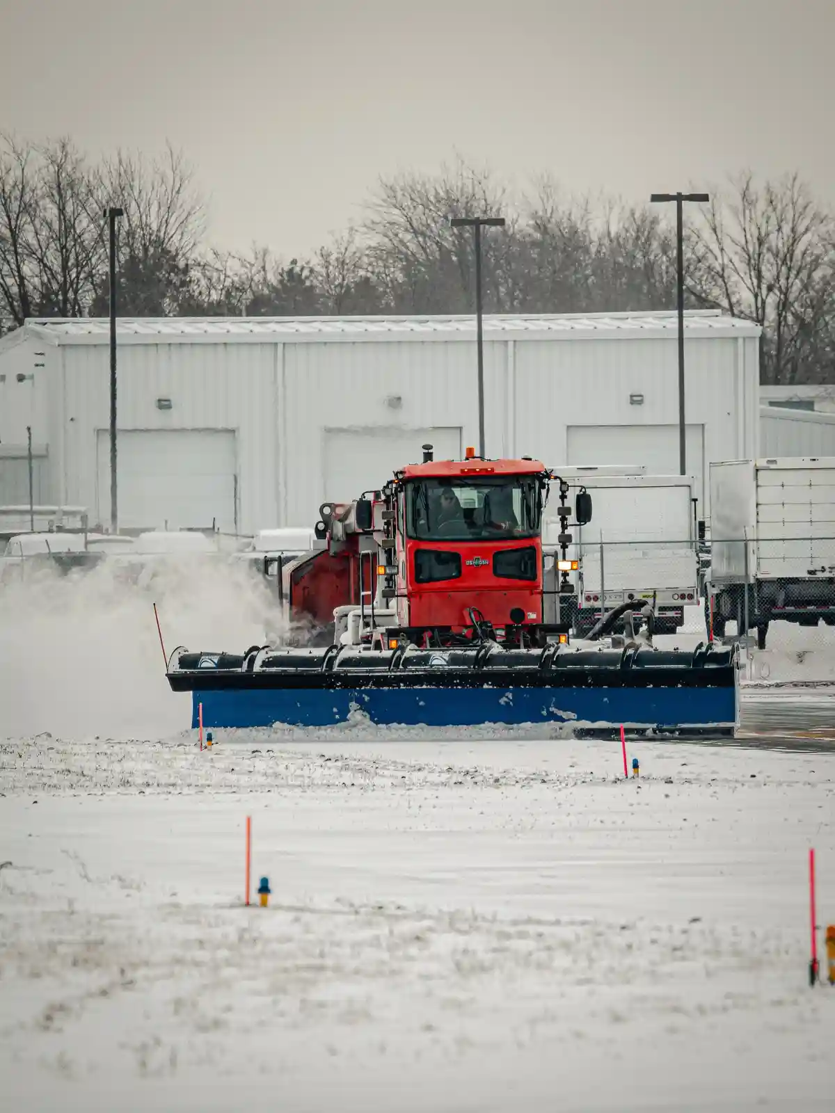 A red snowplow with a large blue blade is actively clearing snow in a snowy industrial area, kicking up a cloud of snow…