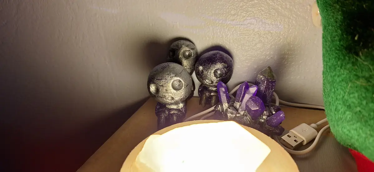 Three small, silver-painted figurines with large eyes sit on a shelf next to a purple crystal cluster and a USB cable…
