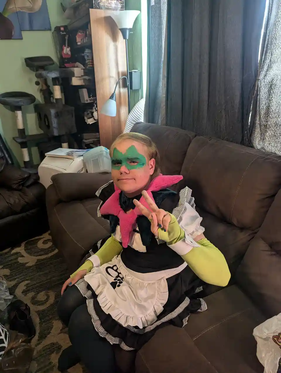 A person sits on a brown couch in a living room, wearing a maid outfit with green face paint and a pink star-shaped collar…